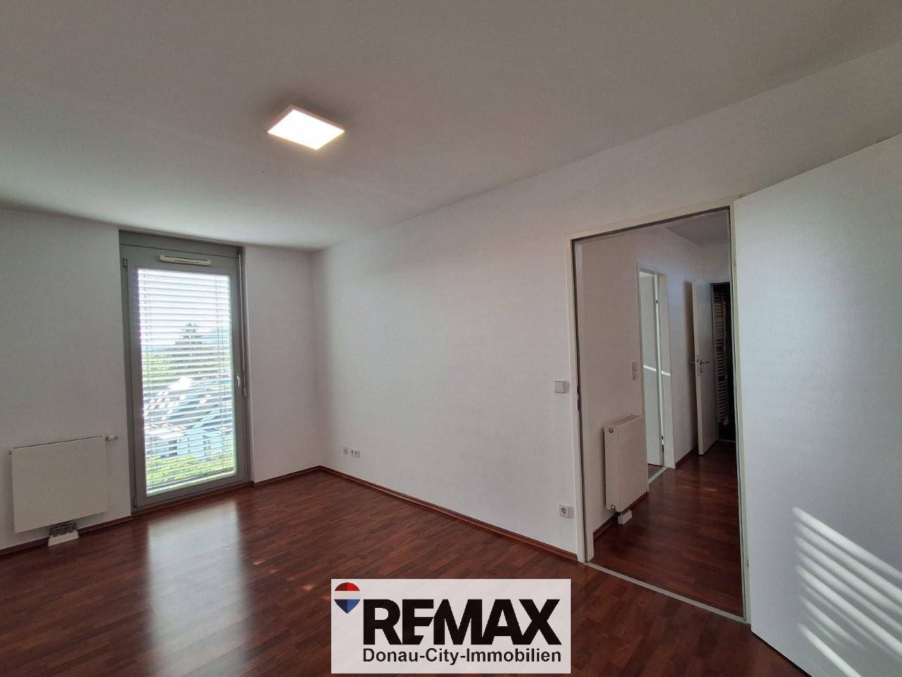 Helles, leeres Zimmer mit dunklem Laminatboden, weißen Wänden und einem hohen Fenster.
