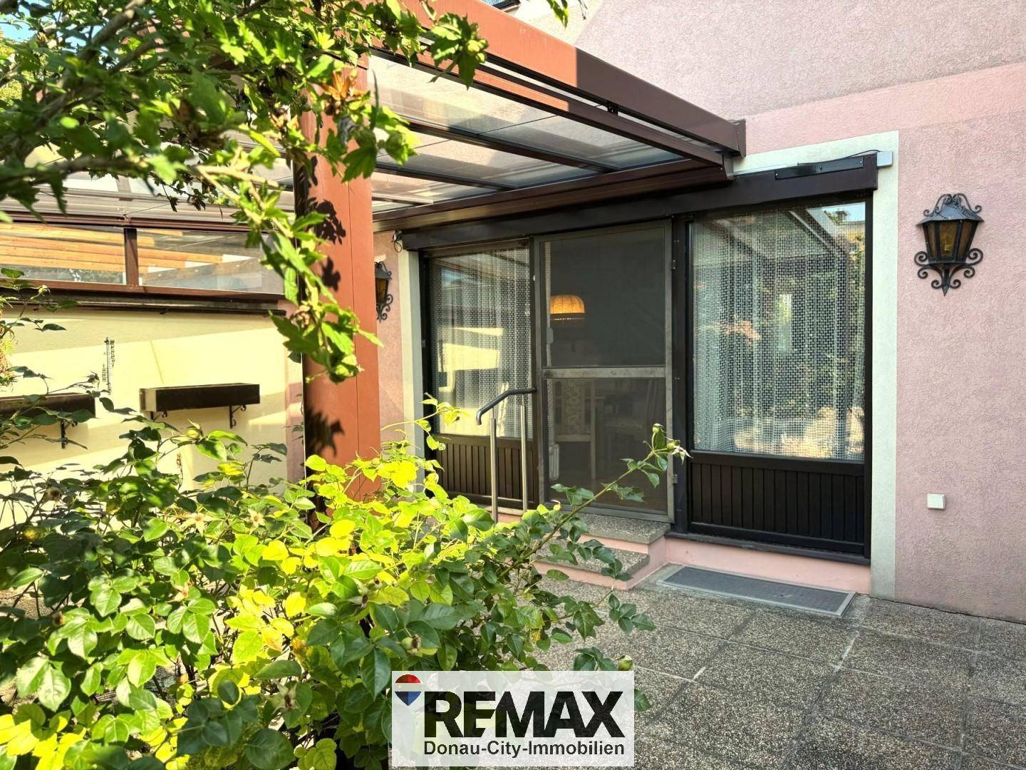 Überdachte Terrasse mit Glasdach und Zugang zum Haus, umgeben von grünen Pflanzen.