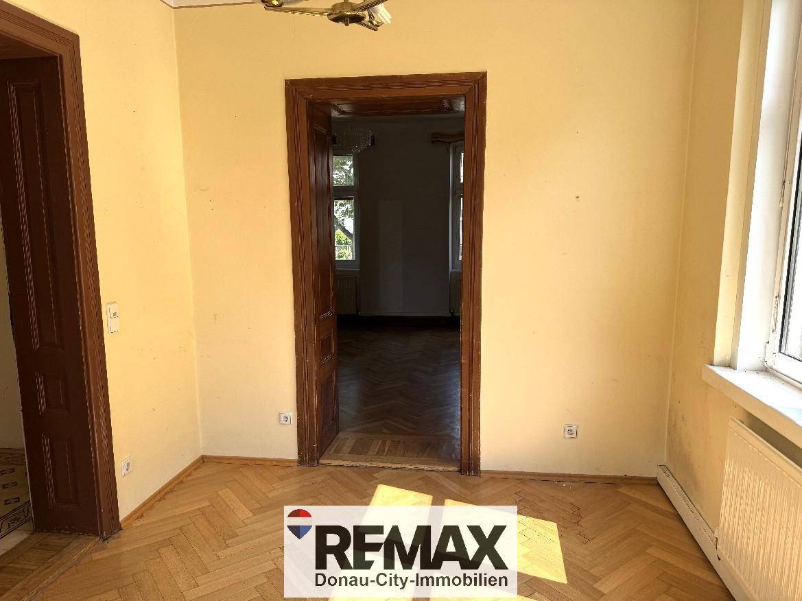 Helles Zimmer mit Fischgrätparkettboden und einer Holztür, die zu einem weiteren Raum führt.