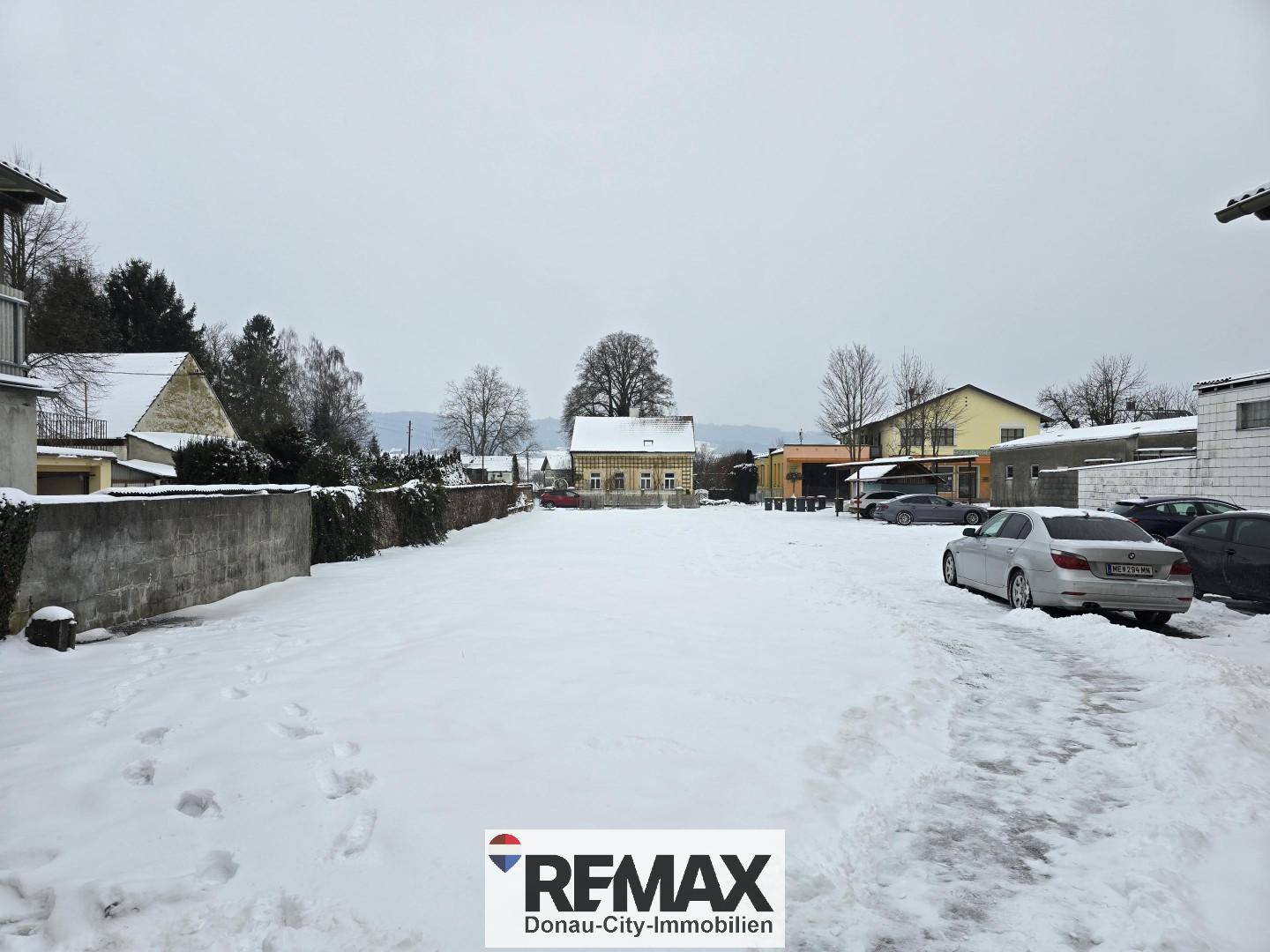 Weitläufige, schneebedeckte Fläche mit Gebäuden und Bäumen im Hintergrund an einem Wintertag.