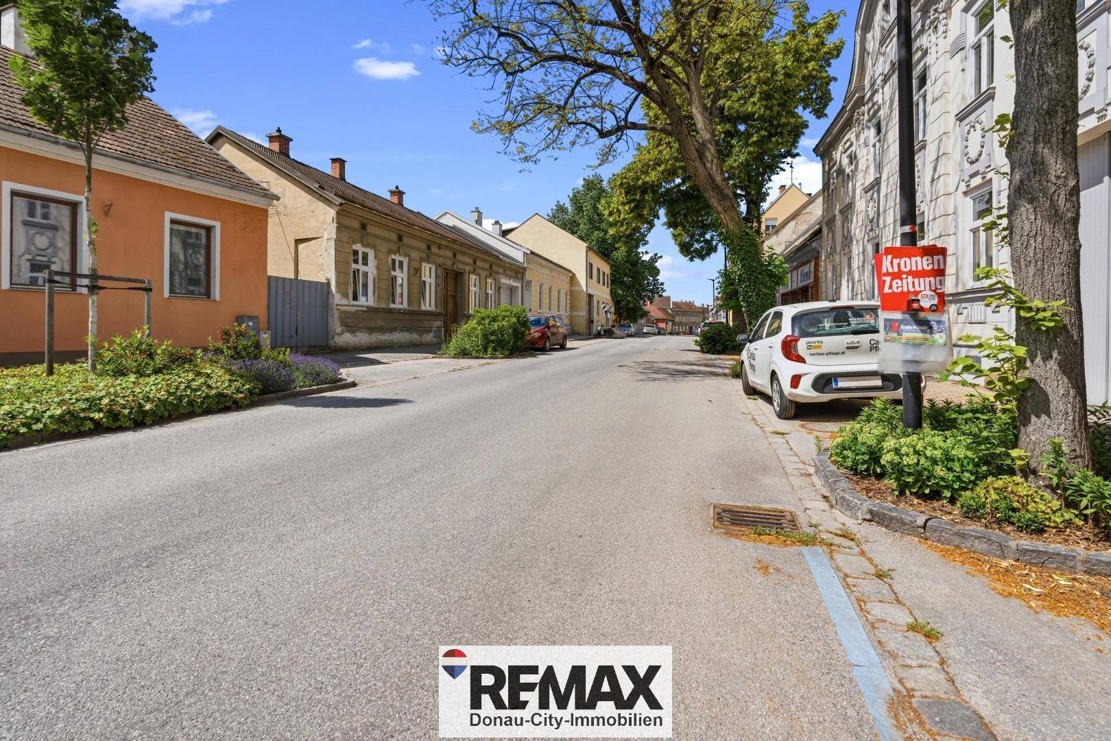 Blick entlang einer ruhigen Wohnstraße mit älteren Gebäuden, Bäumen und parkenden Fahrzeugen.