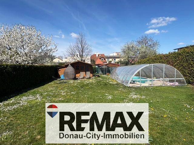 Großer Garten mit Rasenfläche, Gartenhaus und überdachtem Swimmingpool unter blauem Himmel.