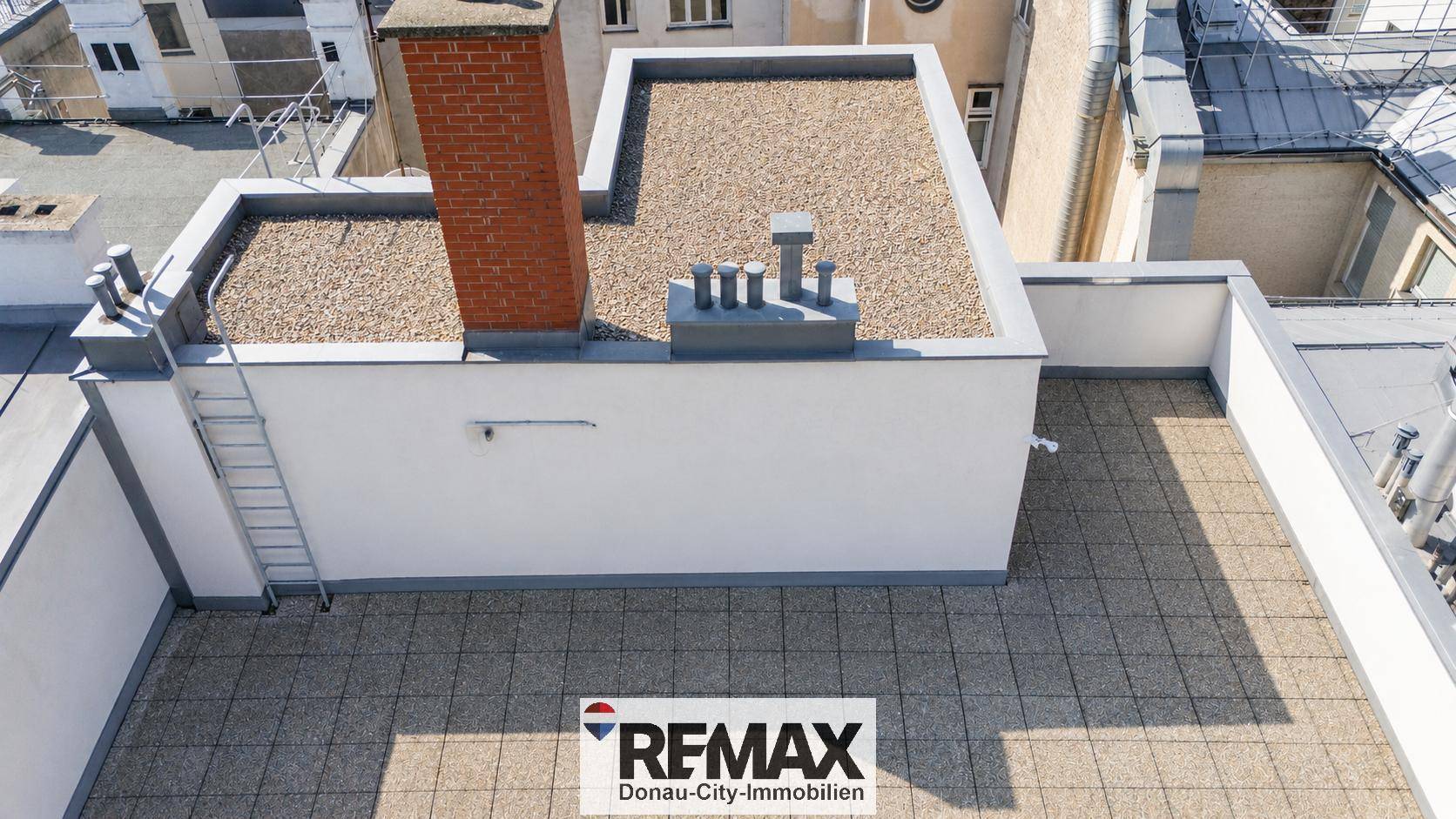 Dachterrasse mit Steinplattenboden und einem Schornstein, umgeben von weiteren Dächern.