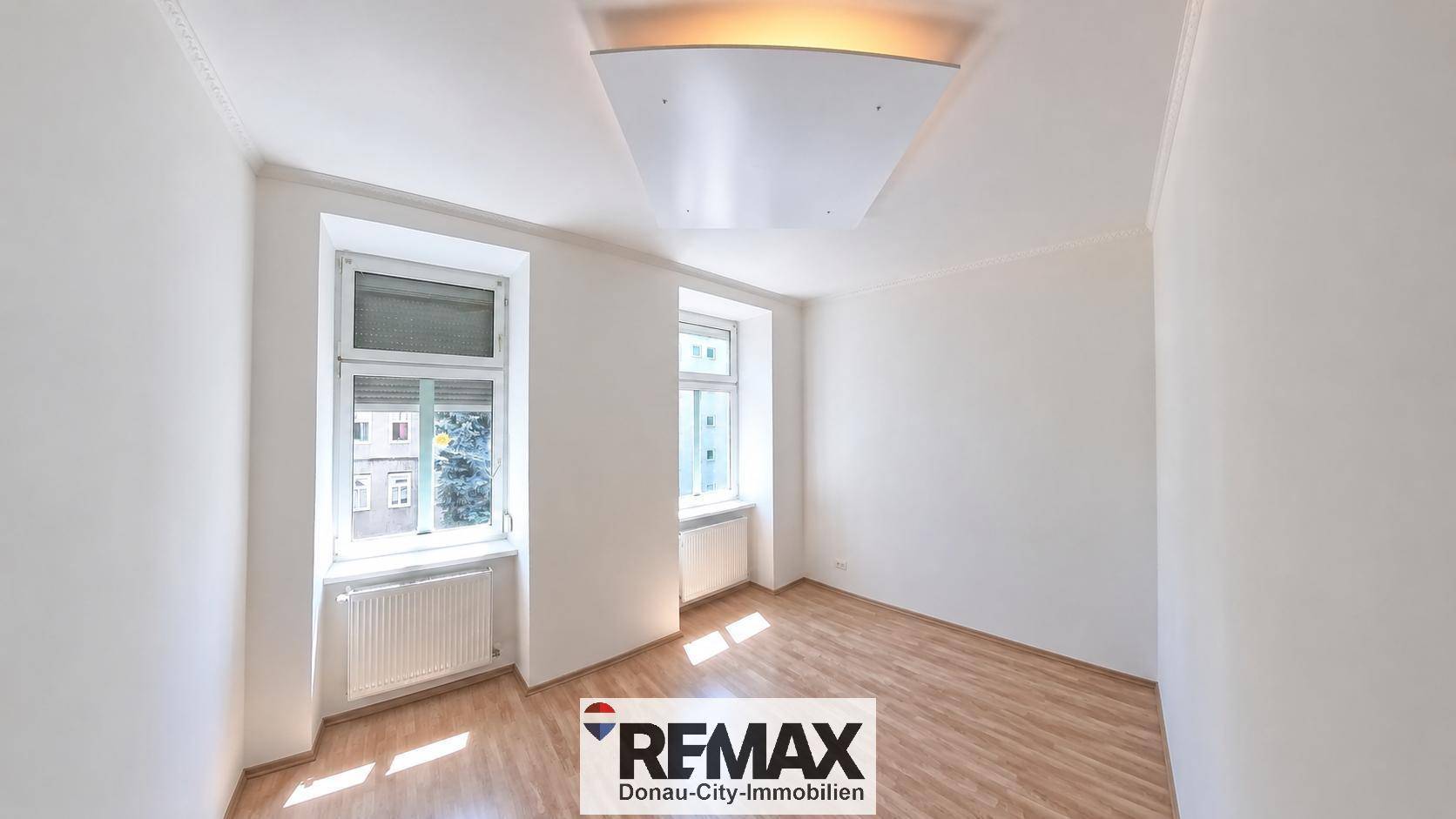 Geräumiges, leeres Zimmer mit zwei Fenstern, Laminatboden und weiß gestrichenen Wänden.