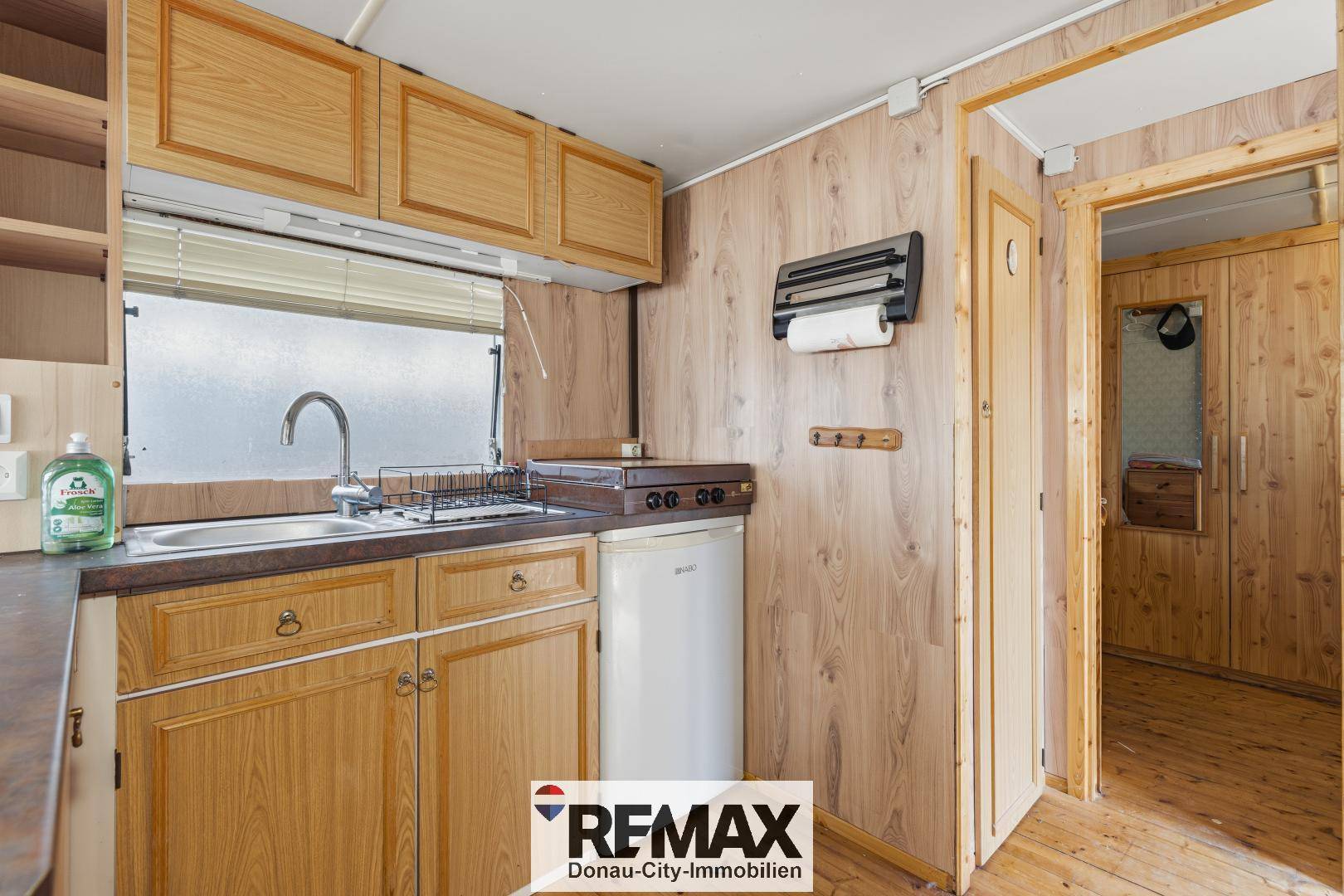 Funktionale Küche mit Holzschränken, Spülbecken und einem kleinen Kühlschrank, angrenzend an einen Flur.