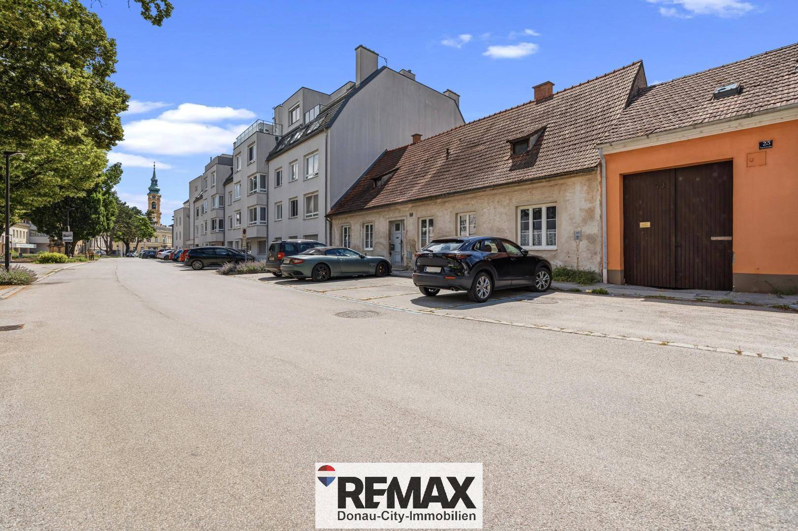 Straßenansicht mit einem älteren Haus und einem modernen Mehrfamilienhaus im Hintergrund sowie parkenden Autos.