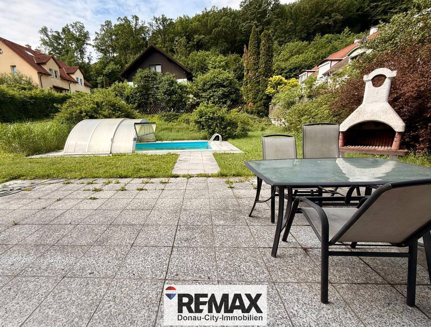 Gepflasterte Terrasse mit Grillplatz und Gartenmöbeln, Blick auf den Pool und die grüne Umgebung.