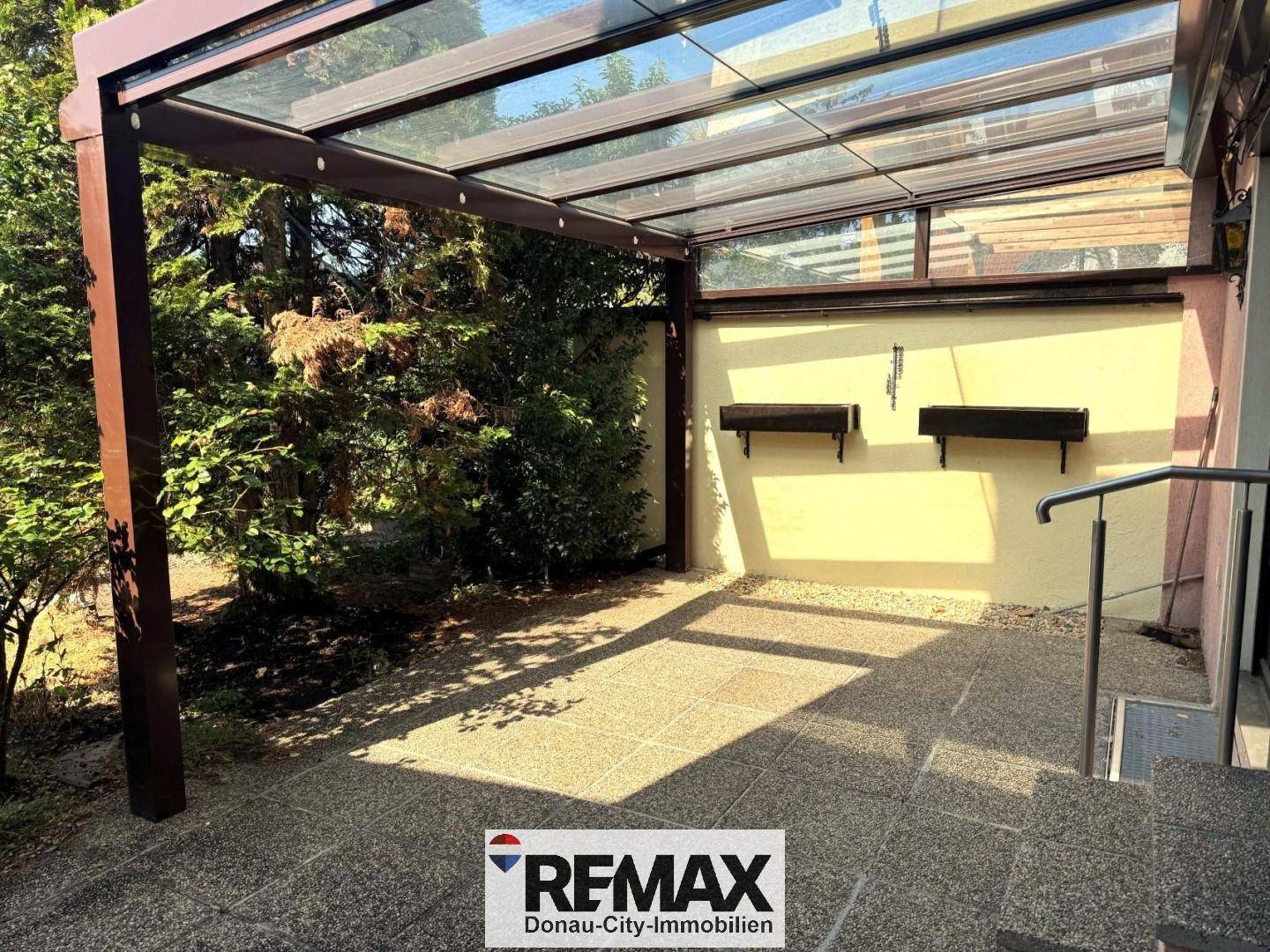 Überdachte Terrasse mit Fliesenboden und Glasdach, umgeben von üppigem Grün.