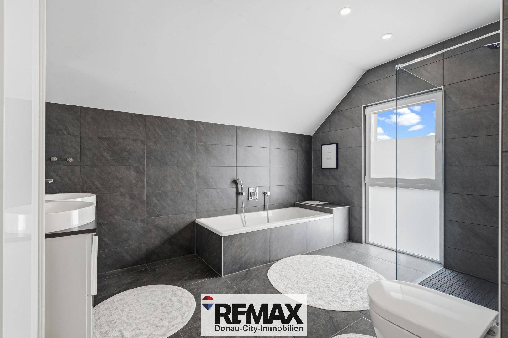 Geräumiges Badezimmer mit Badewanne, Dusche, dunklen Fliesen und Fenster unter der Dachschräge.