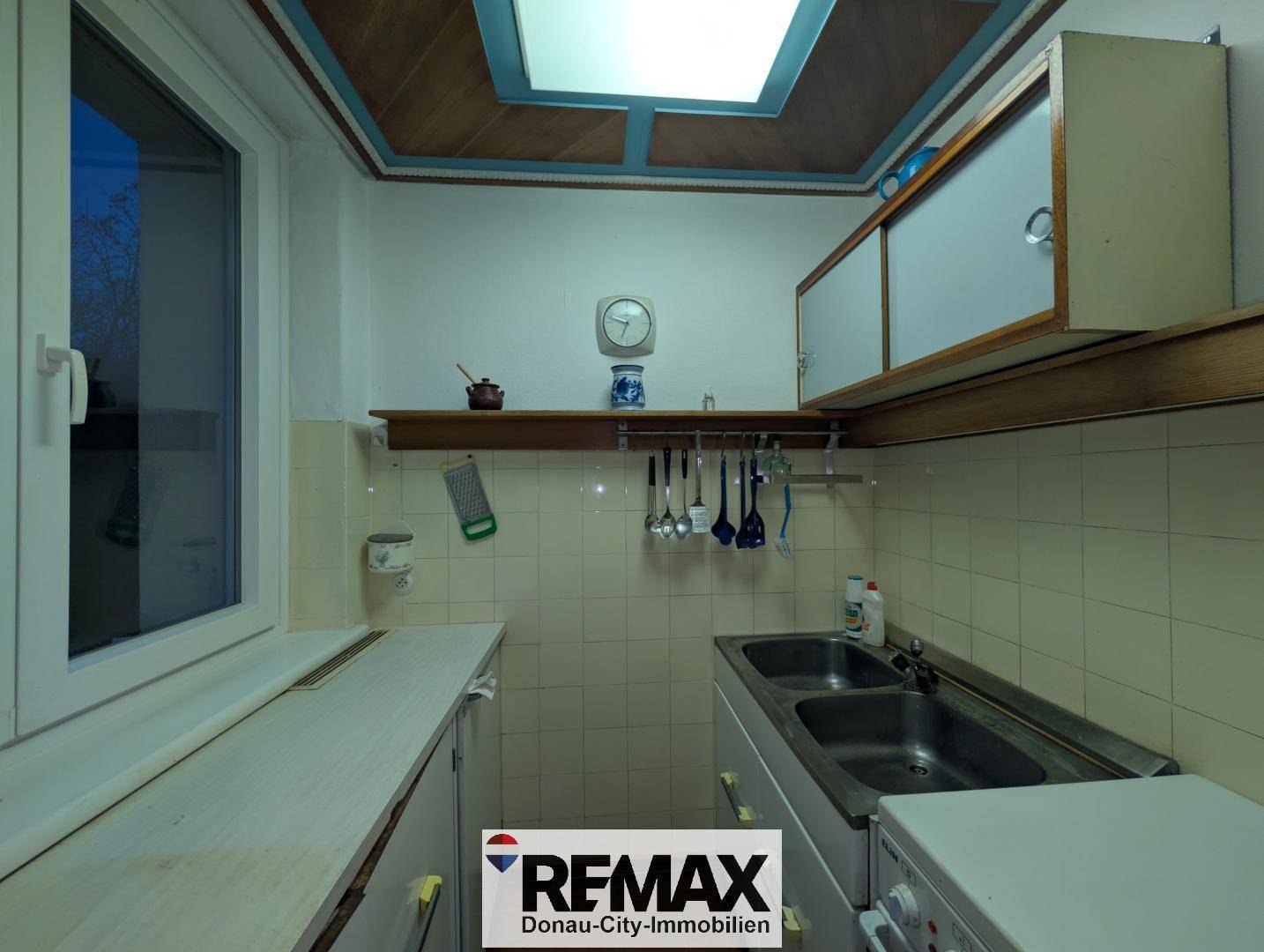 Kleine Küche mit doppelter Spüle, gefliesten Wänden und einem Fenster, das Tageslicht hereinlässt.