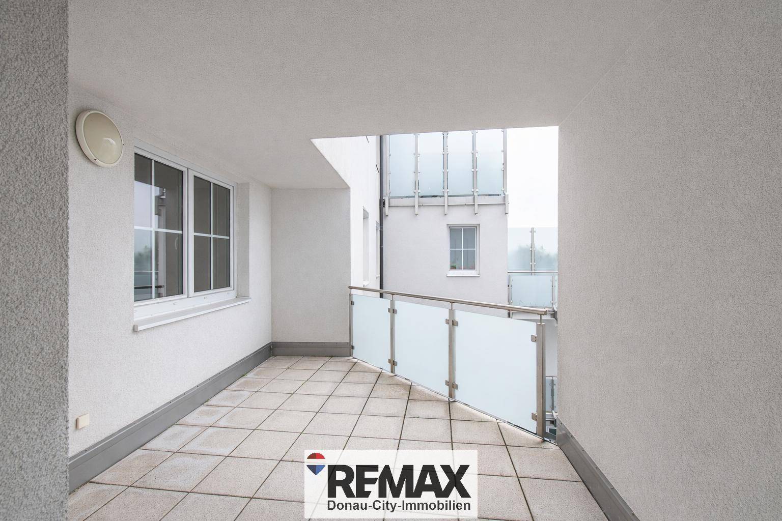 Überdachte Loggia mit Fliesenboden und moderner Glasbrüstung, bietet einen geschützten Außenbereich.