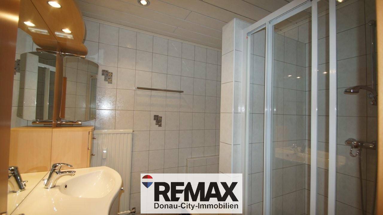 Badezimmer mit Waschbecken, Spiegel, Heizkörper und einer Duschkabine, hell gefliest.