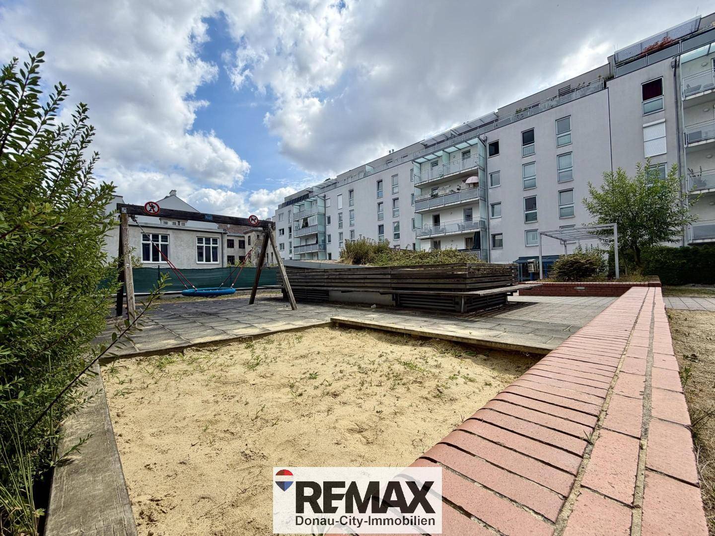Gemeinschaftlicher Spielplatz mit Sandkasten und Schaukel, umgeben von Wohngebäuden.