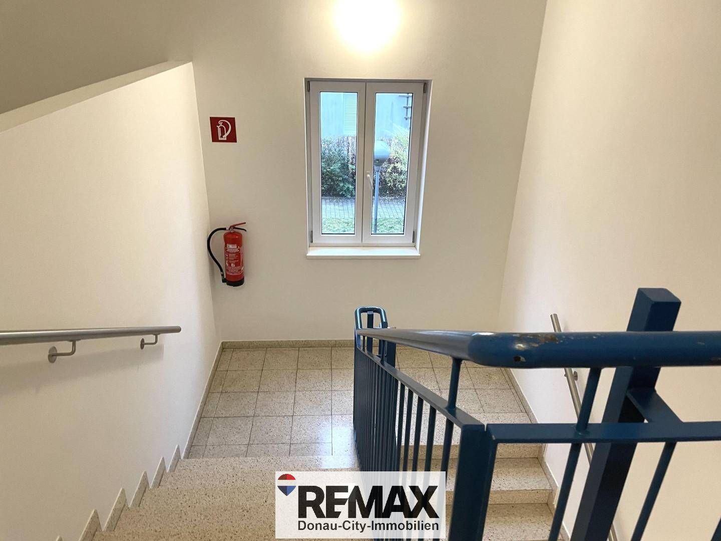 Treppenhaus mit Fliesenboden, Geländer und Fenster zum Außenbereich.