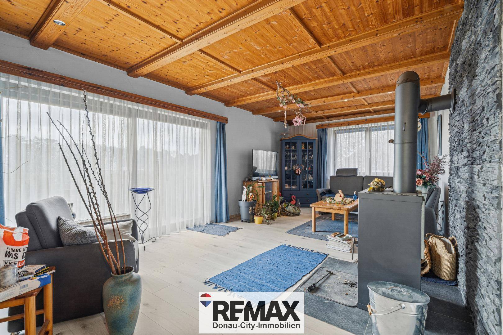 Helles Wohnzimmer mit großen Fenstern, Holzbalkendecke und einem Kaminofen.