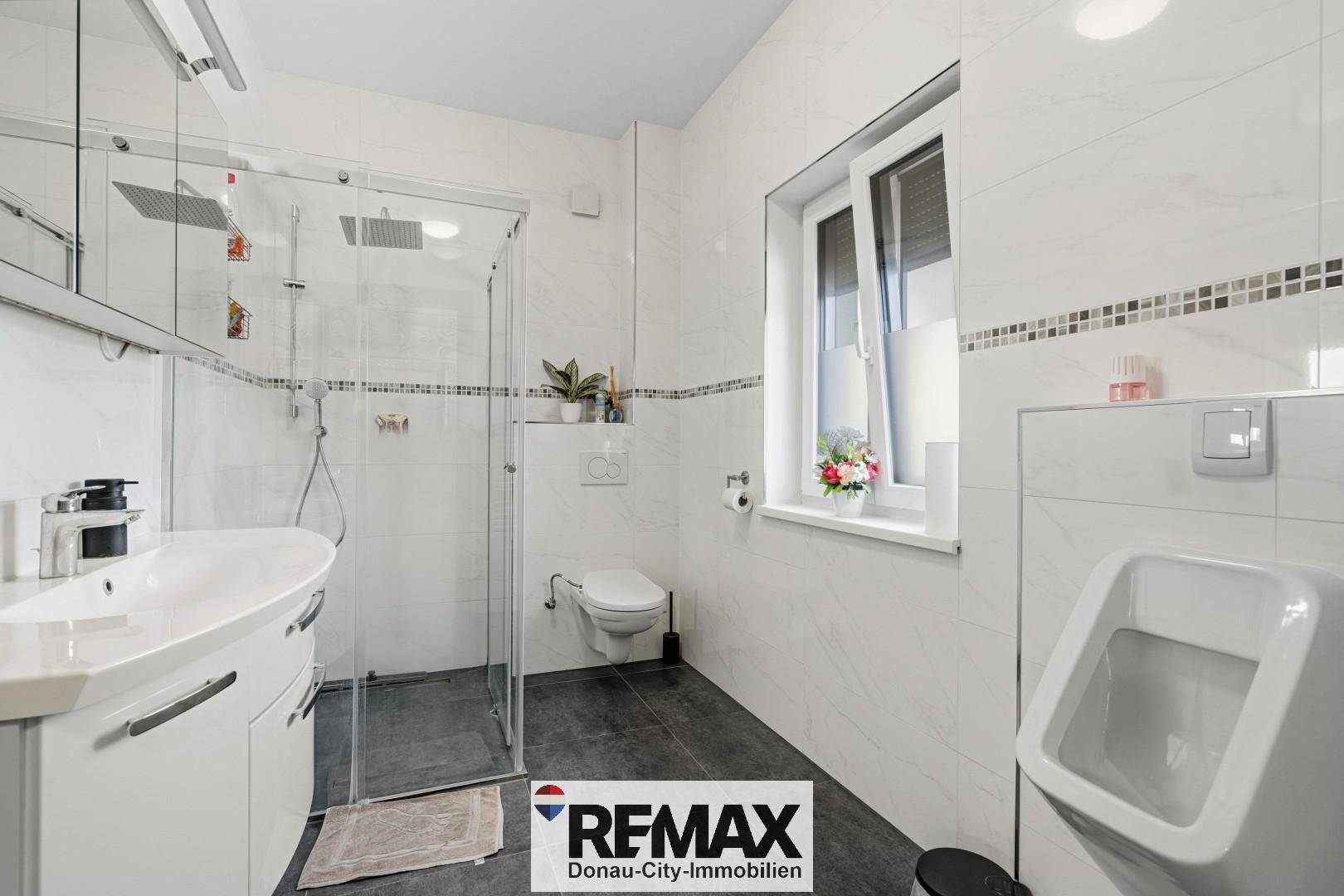 Modernes Badezimmer mit Dusche, Urinal, WC und weißen Fliesen mit Akzentstreifen.