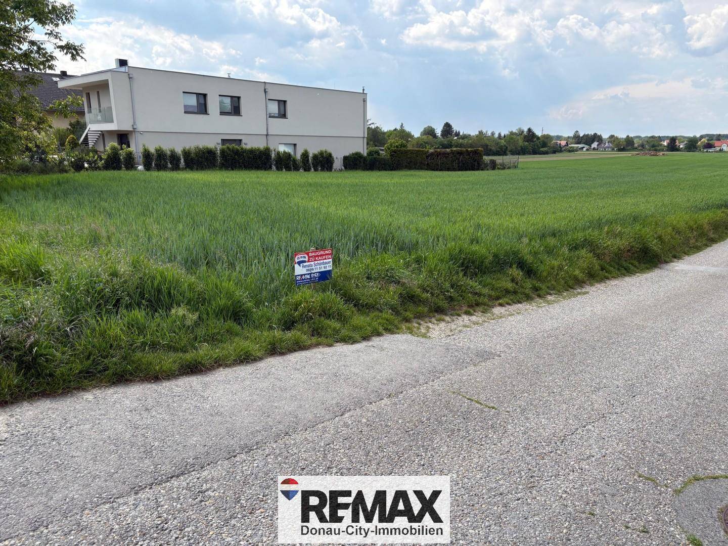 Grünes Feld mit einem modernen Gebäude im Hintergrund und einem 'Zu Verkaufen'-Schild am Wegrand.