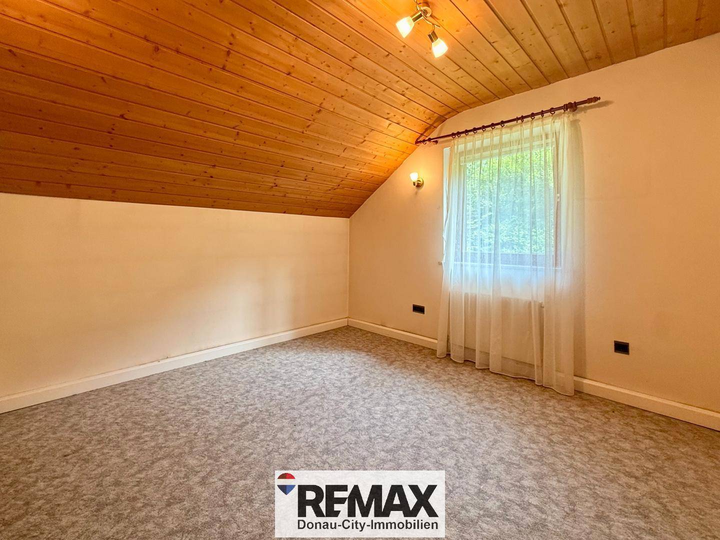 Schlafzimmer im Dachgeschoss mit schräger Holzdecke, Teppichboden und Fenster mit Blick ins Grüne.