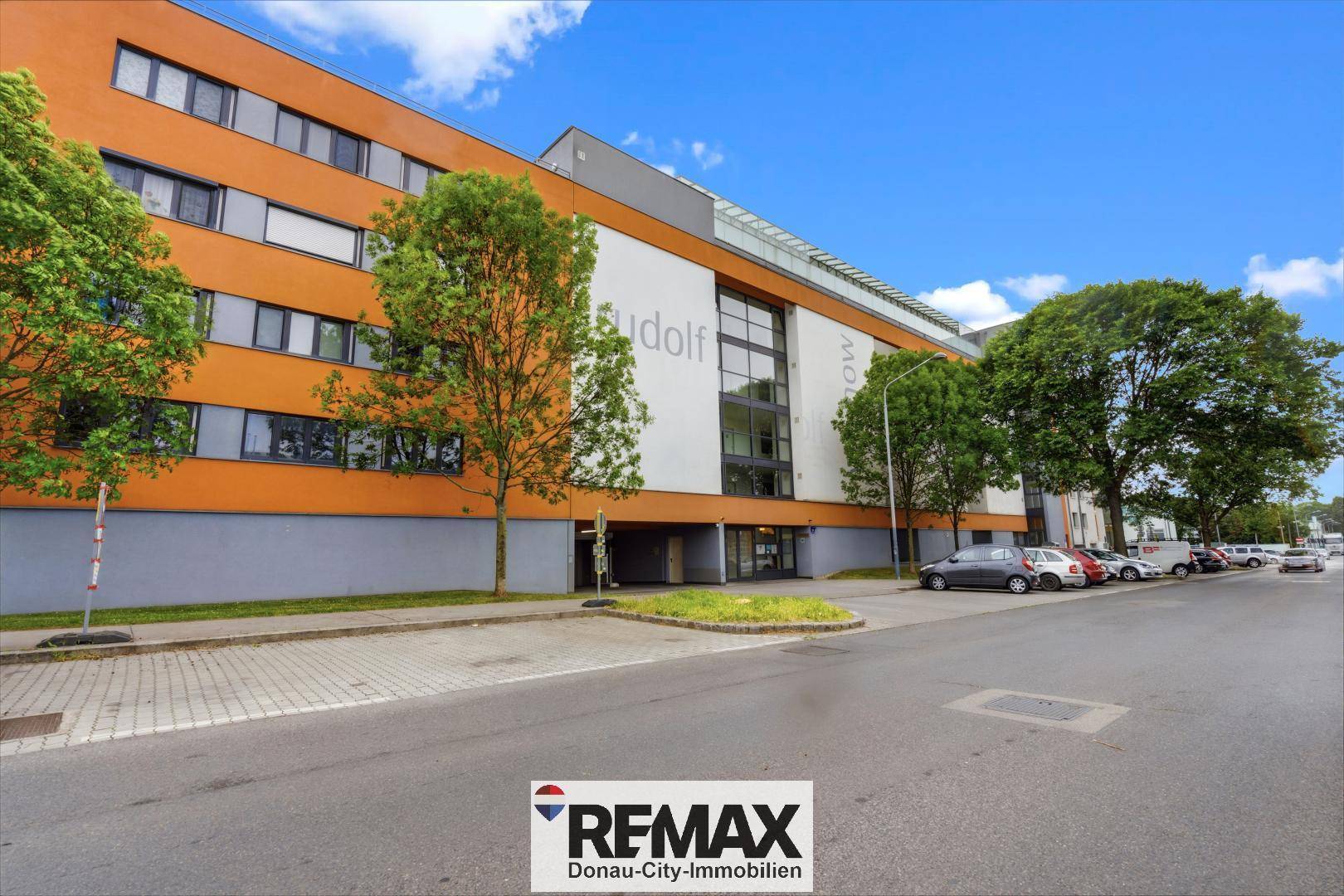 Moderne Gebäudefassade mit orangefarbenen Akzenten und großen Fenstern an einer Straße.