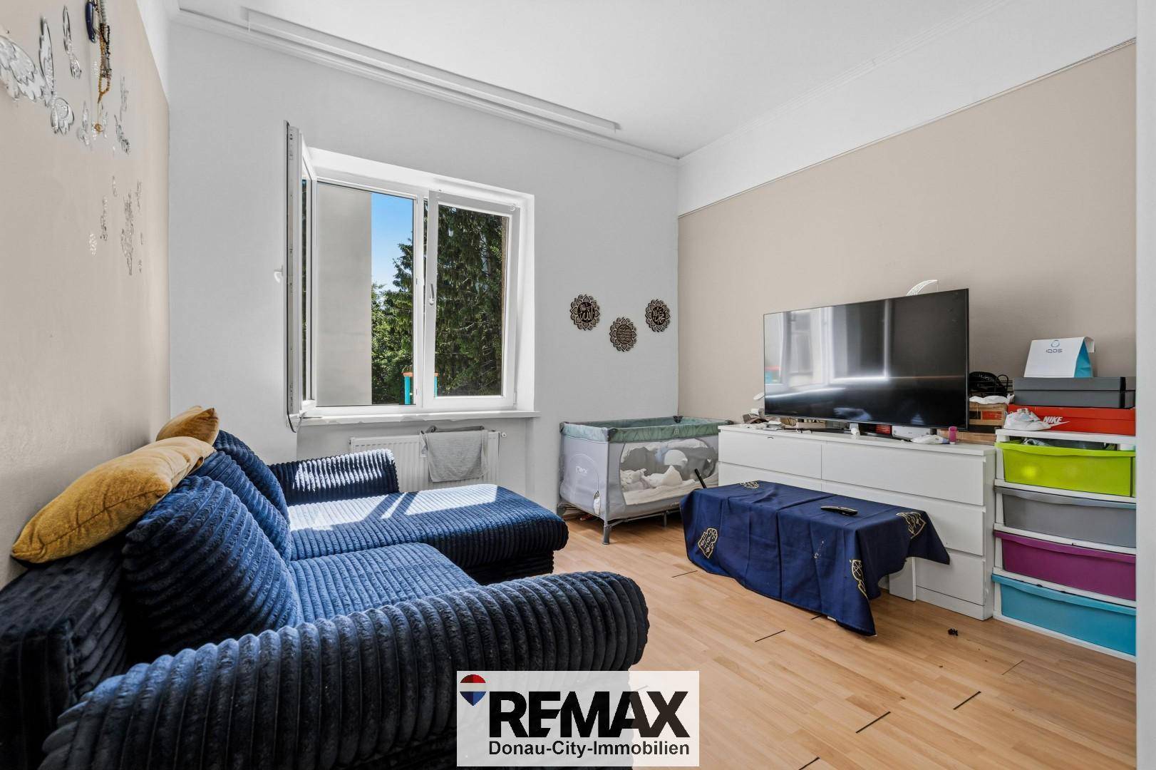 Wohnzimmer mit Fernseher, Babybett und Blick ins Grüne durch das offene Fenster.