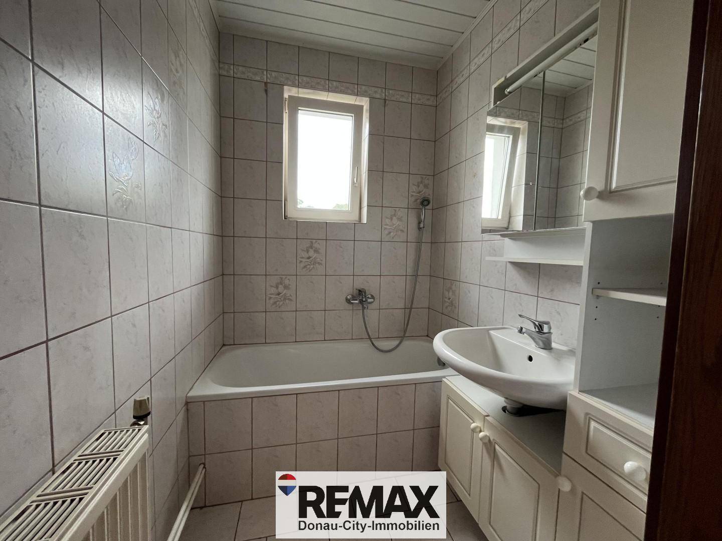 Helles Badezimmer mit Badewanne, Waschbecken, Fenster und weißen Fliesen im 80er-Jahre-Stil.