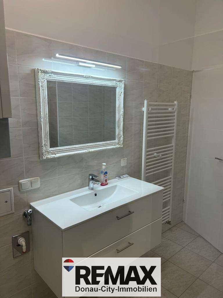 Badezimmer mit Waschtisch, Spiegel und Heizkörper, alles in hellen Grautönen gehalten.