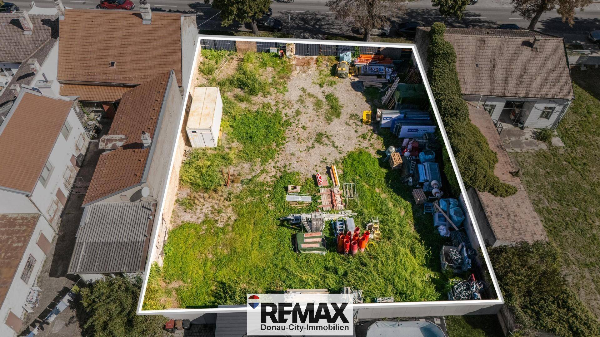 Umrandetes Grundstück mit hohem Gras, Baumaterialien und angrenzenden Gebäuden.