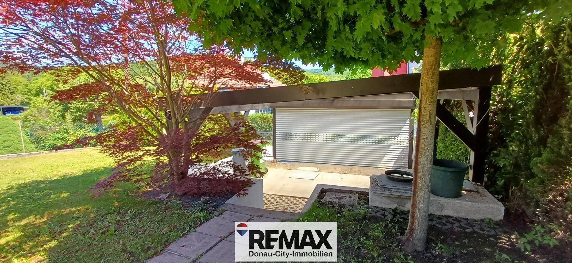 Überdachter Bereich im Garten, möglicherweise ein Carport oder Abstellplatz, umgeben von Bäumen.