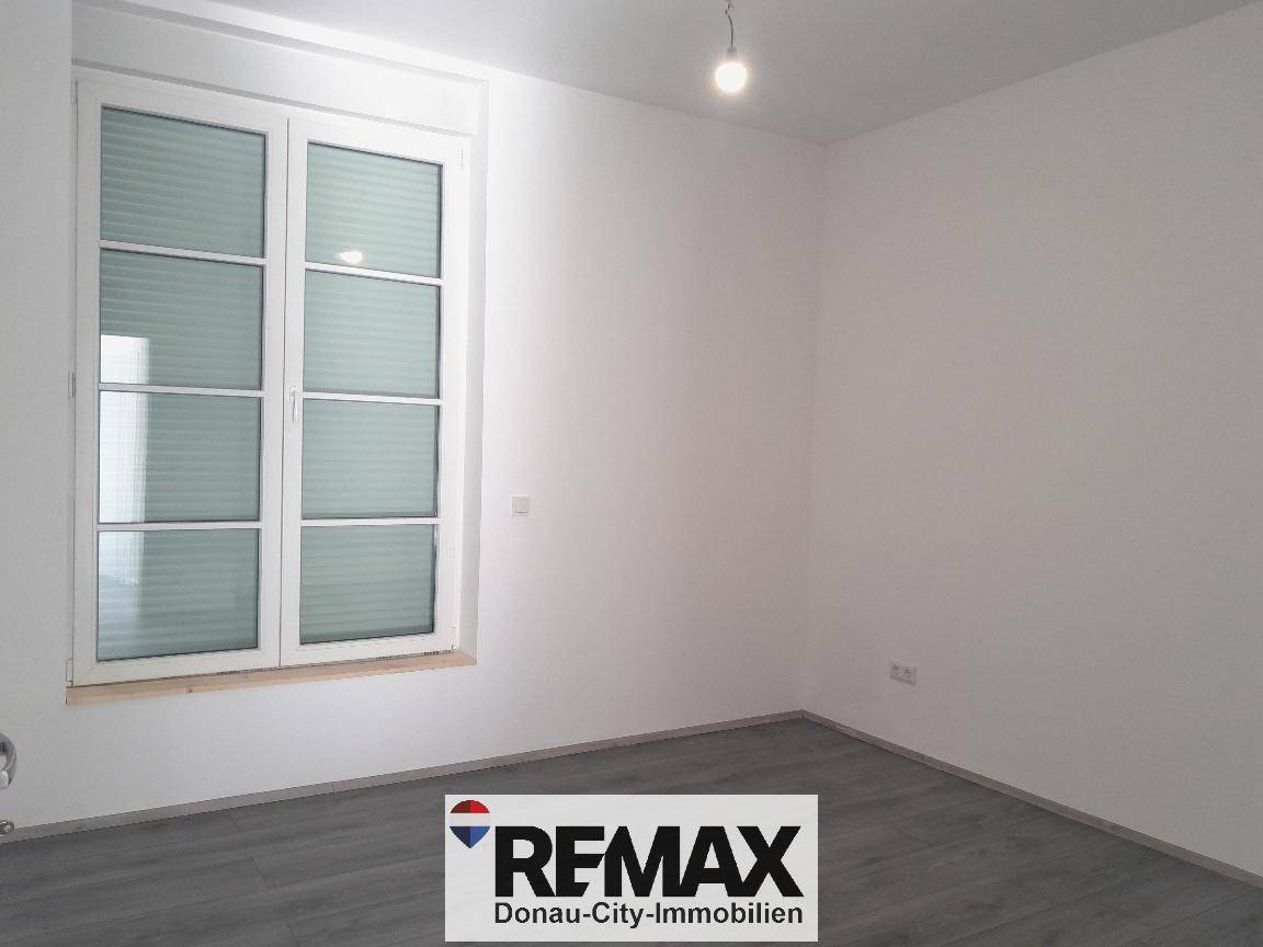 Heller, leerer Raum mit großem Fenster und modernem Laminatboden.