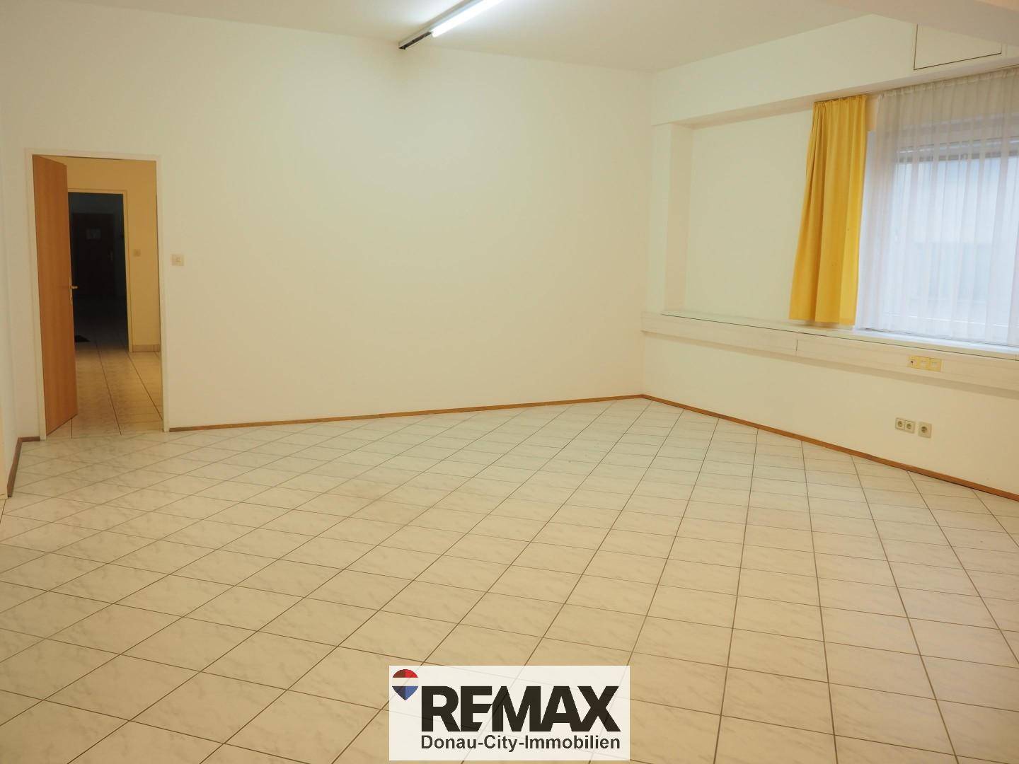 Heller Büroraum mit Fliesenboden, weißen Wänden und einem Fenster mit gelben Vorhängen und Türzugang.