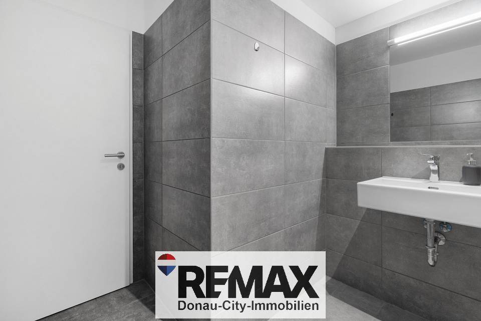 Modernes Badezimmer mit grauen Fliesen, wandhängendem Waschbecken und weißer Tür.