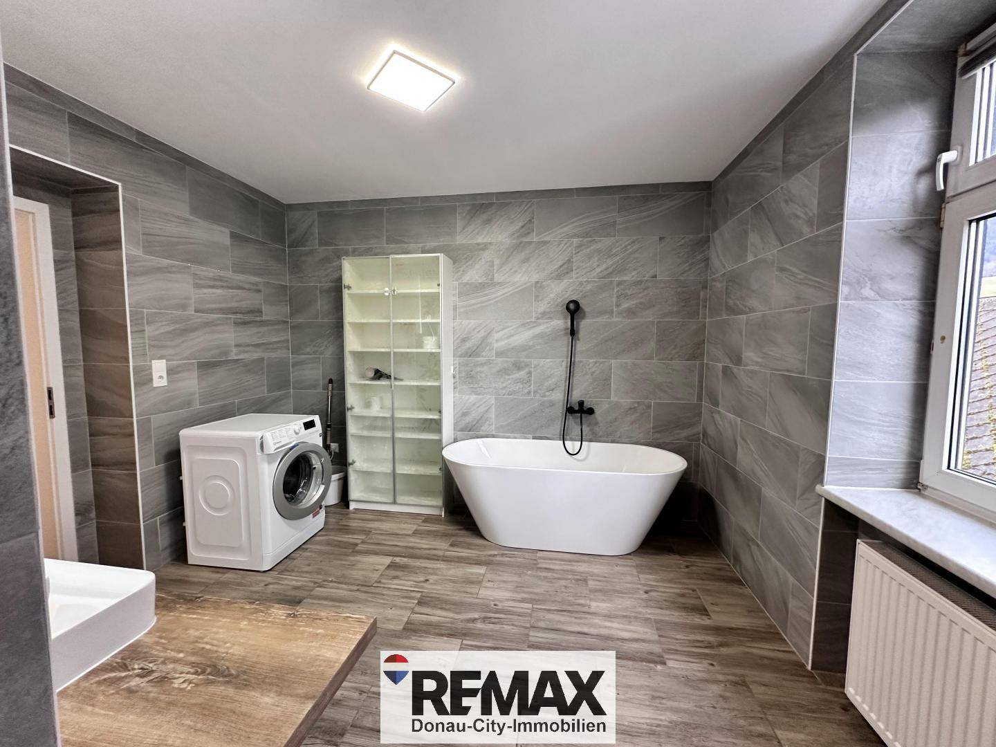 Geräumiges Badezimmer mit modernen grauen Fliesen, freistehender Badewanne und Waschmaschine.