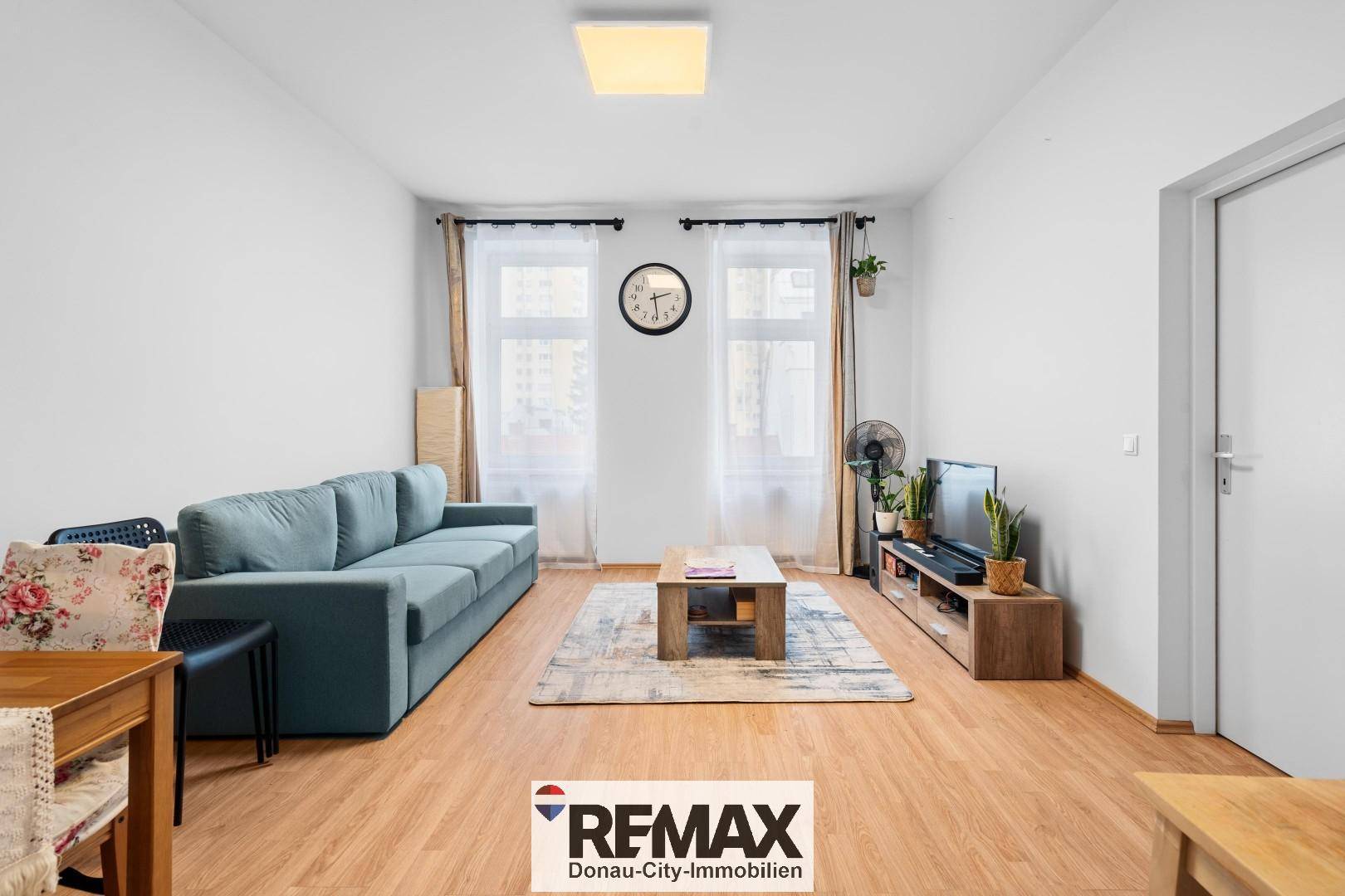 Gemütliches Wohnzimmer mit hellem Laminatboden, blauem Sofa und zwei großen Fenstern.