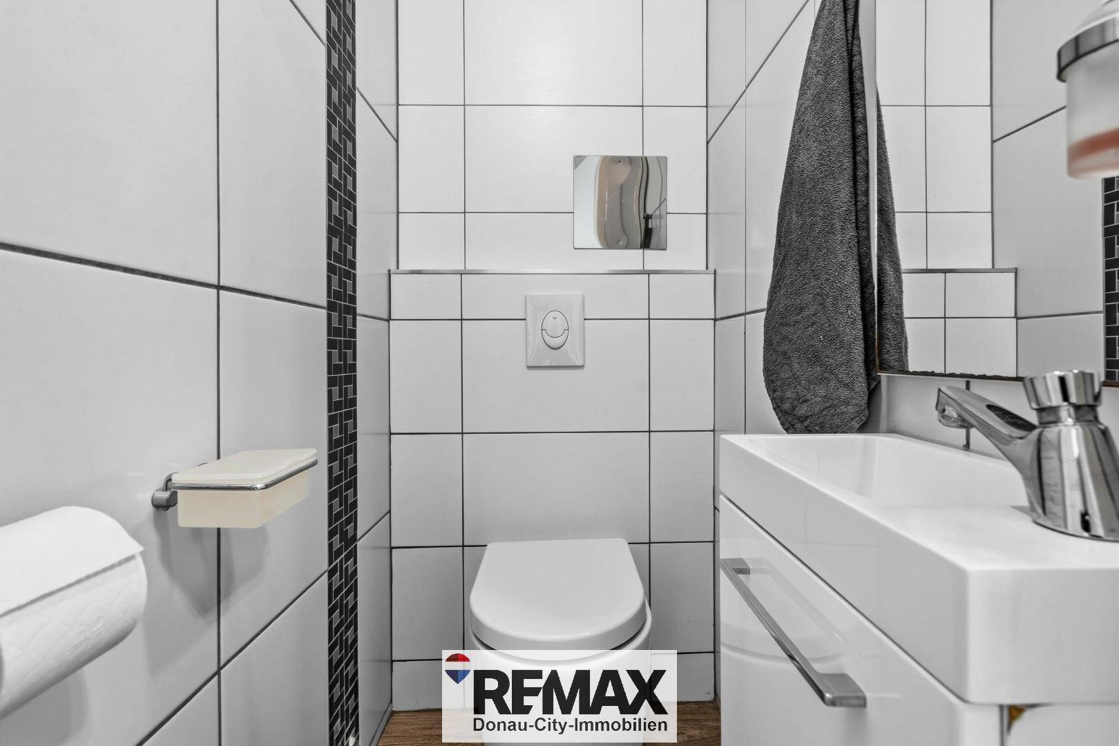 Separates WC mit wandhängender Toilette und kleinem Waschbecken, komplett gefliest in Weiß.