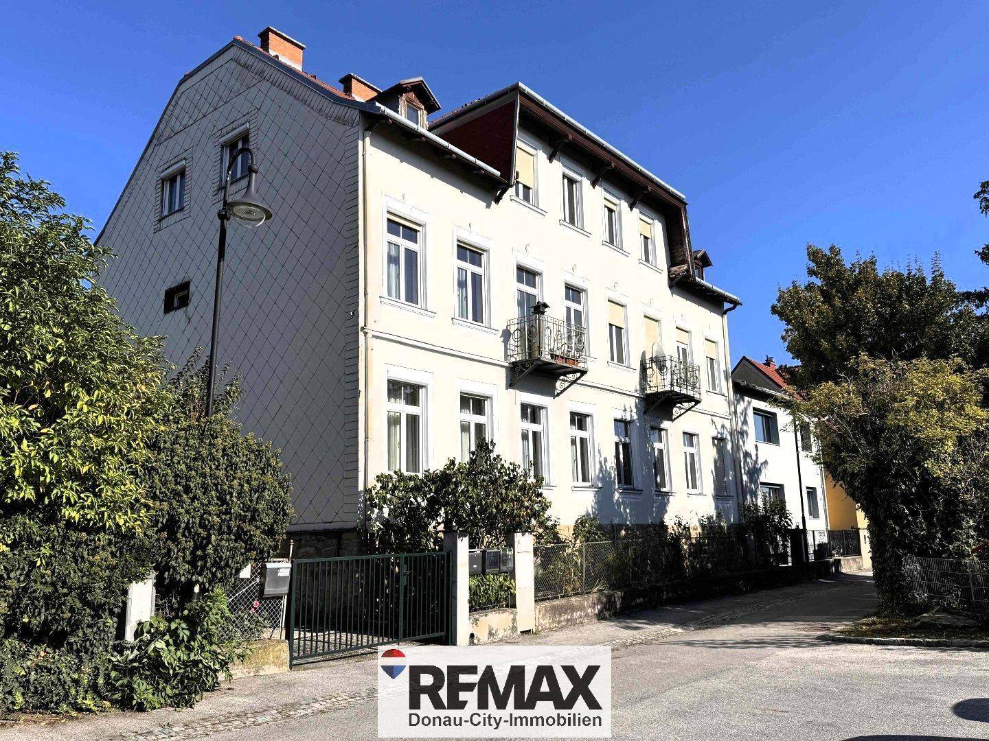 Historisches Mehrfamilienhaus mit klassischer Fassade, kleinen Balkonen und grüner Umgebung an einer Straße.