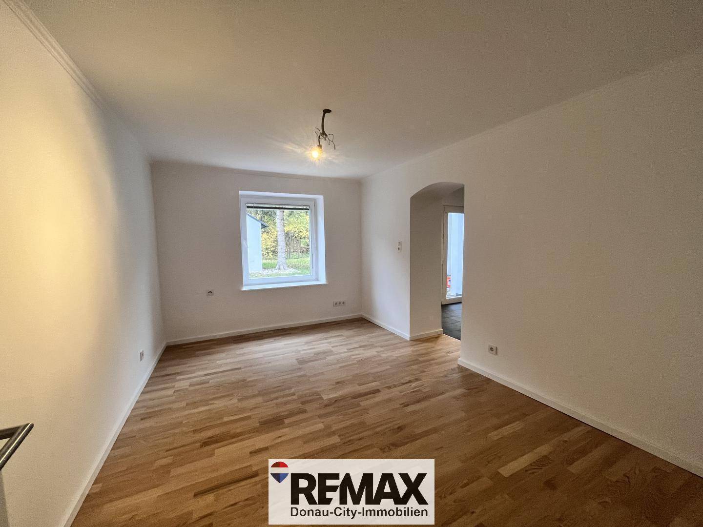 Helles Wohnzimmer mit Parkettboden, weiß gestrichenen Wänden und Fensterblick ins Grüne.