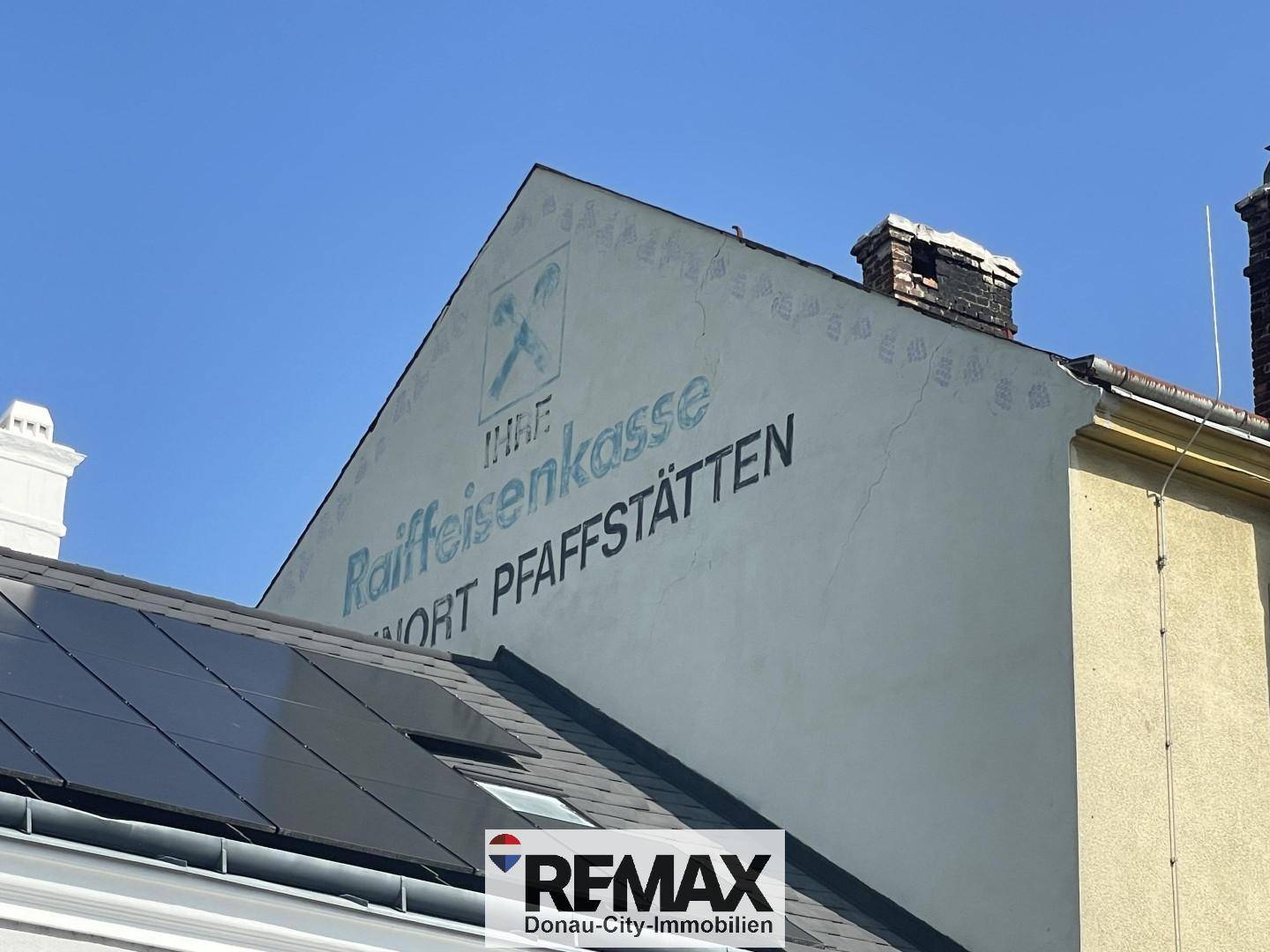 Nahaufnahme einer Gebäudewand mit alter Raiffeisenkasse-Beschriftung und Solarpaneelen auf dem Dach.