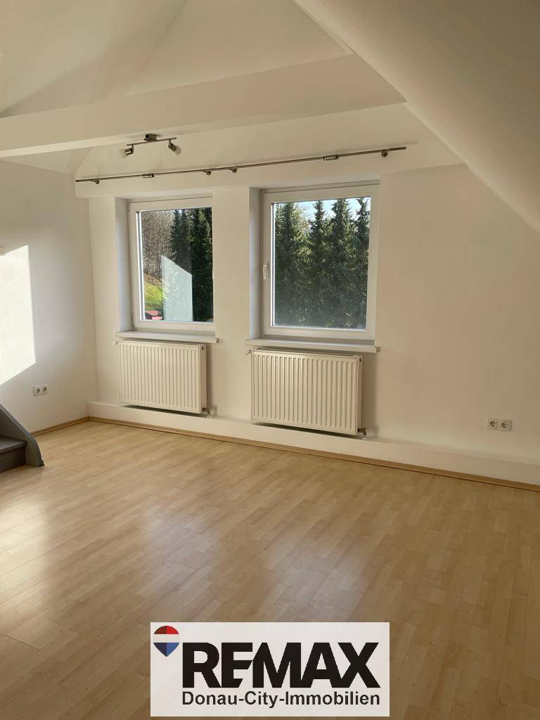 Lichtdurchfluteter Raum mit Holzfußboden und zwei Fenstern, die einen Blick ins Grüne bieten.