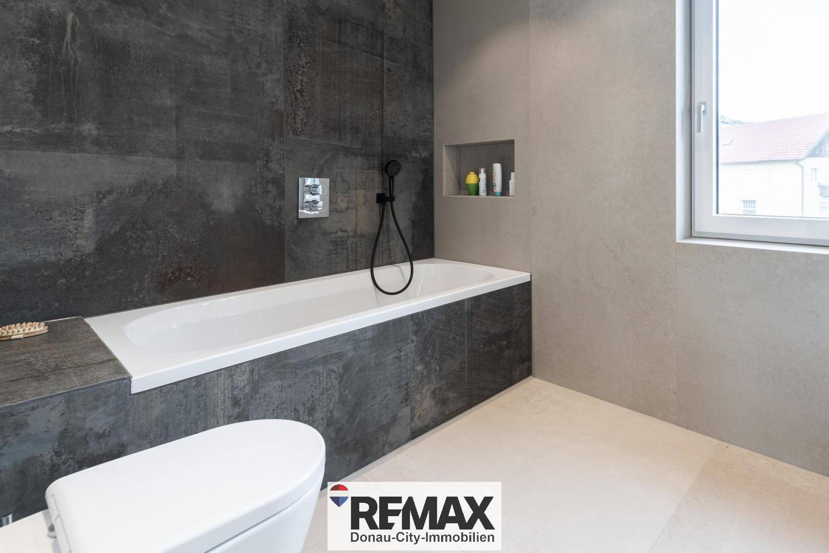 Modernes Badezimmer mit Badewanne, dunklen Fliesen und Fenster für Tageslicht.