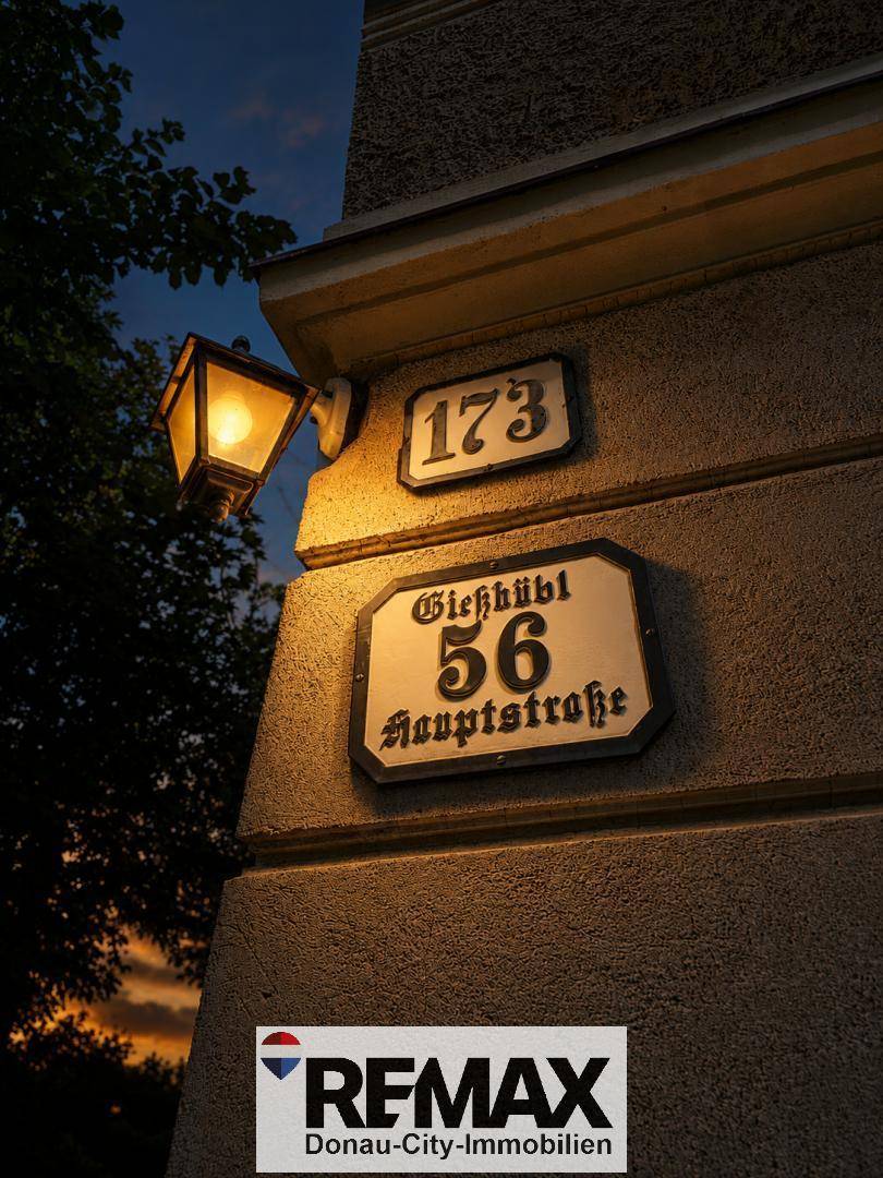 Hausnummern und Straßenschilder an einer Fassade, beleuchtet von einer historischen Laterne in der Dämmerung.