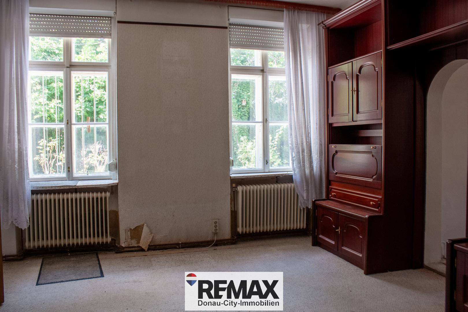 Renovierungsbedürftiges Zimmer mit zwei Fenstern, Heizkörpern und einem großen Holzschrank.