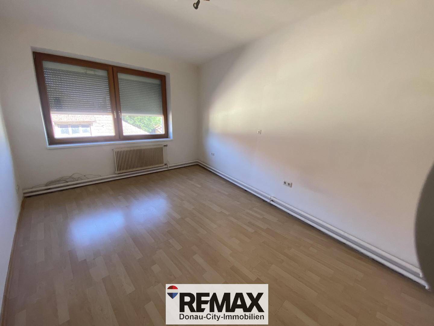Leeres Schlafzimmer mit hellem Laminatboden, Fenster mit Rollläden und weiß gestrichenen Wänden.