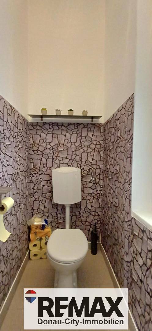 Kleines Gäste-WC mit gemusterter Tapete, Toilette und einem kleinen Regal über der Spülung.
