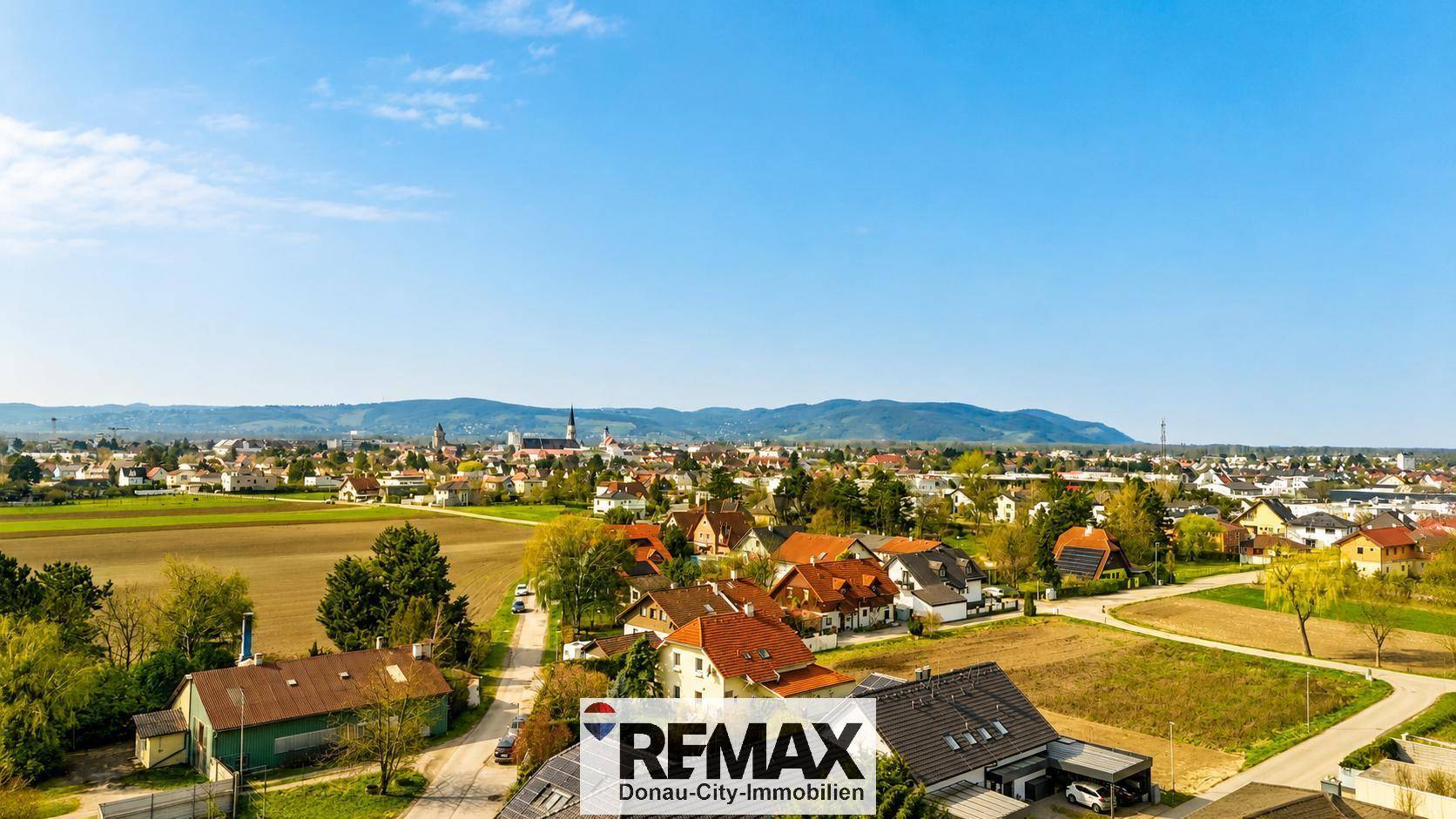 Panoramablick über eine Stadt mit Wohngebäuden, Feldern und Bergen im Hintergrund.