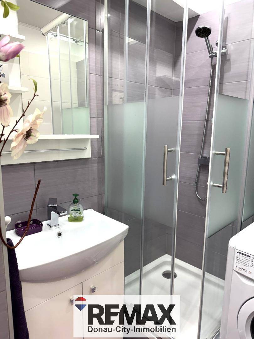 Modernes Badezimmer mit Duschkabine, Waschbecken und Waschmaschine in grauen Fliesen.