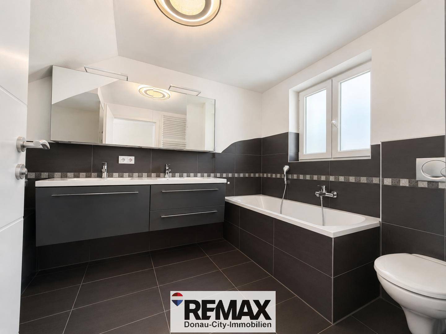 Modernes Badezimmer mit Doppelwaschbecken, Badewanne und dunklen Fliesen.