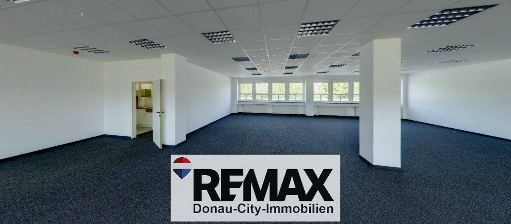 Geräumiges, leeres Büro mit dunklem Teppichboden, vielen Fenstern und Zugang zu einer kleinen Küche.