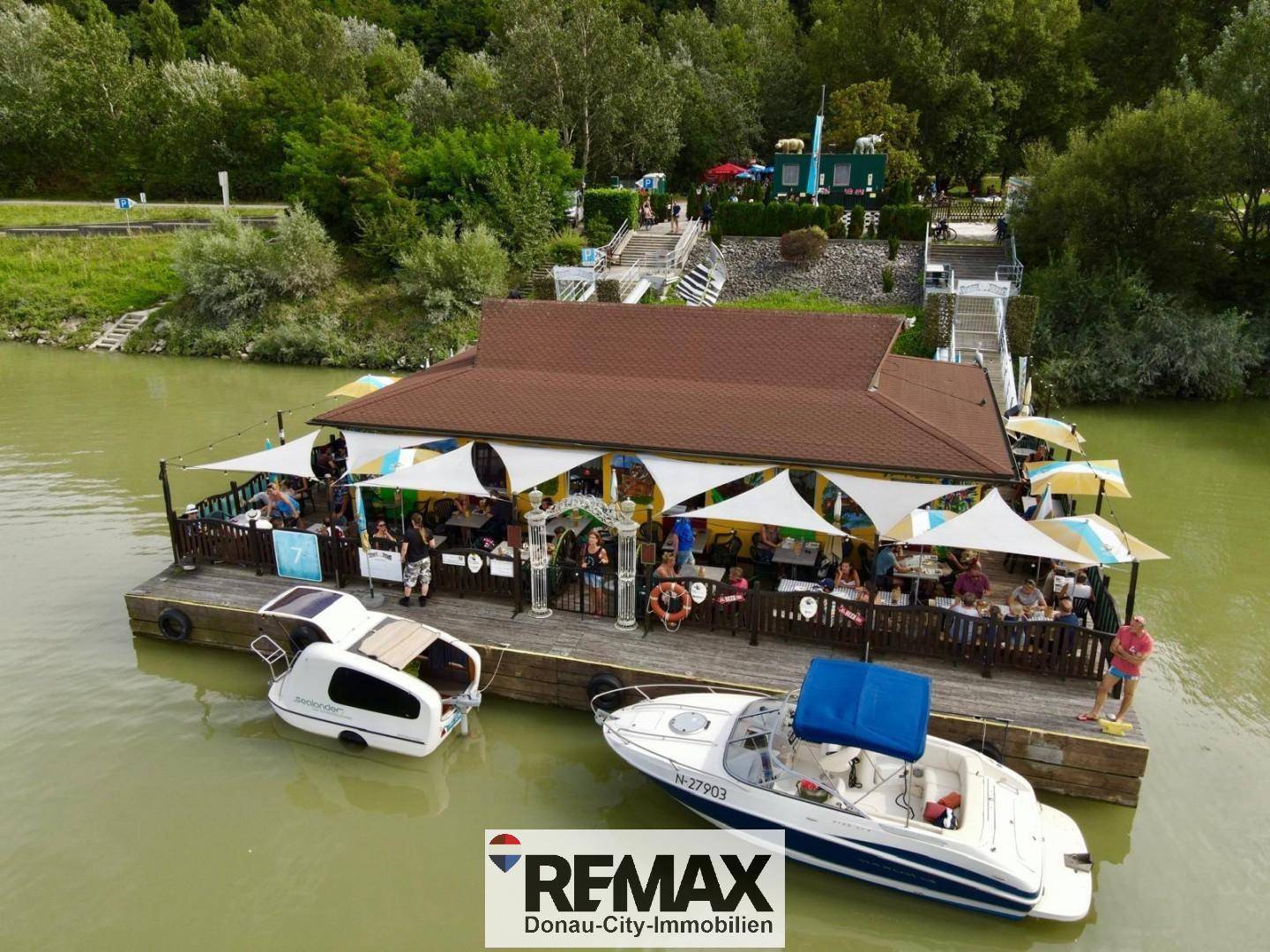 Luftaufnahme des schwimmenden Restaurants mit belebter Außenterrasse und angedockten Booten am Flussufer.