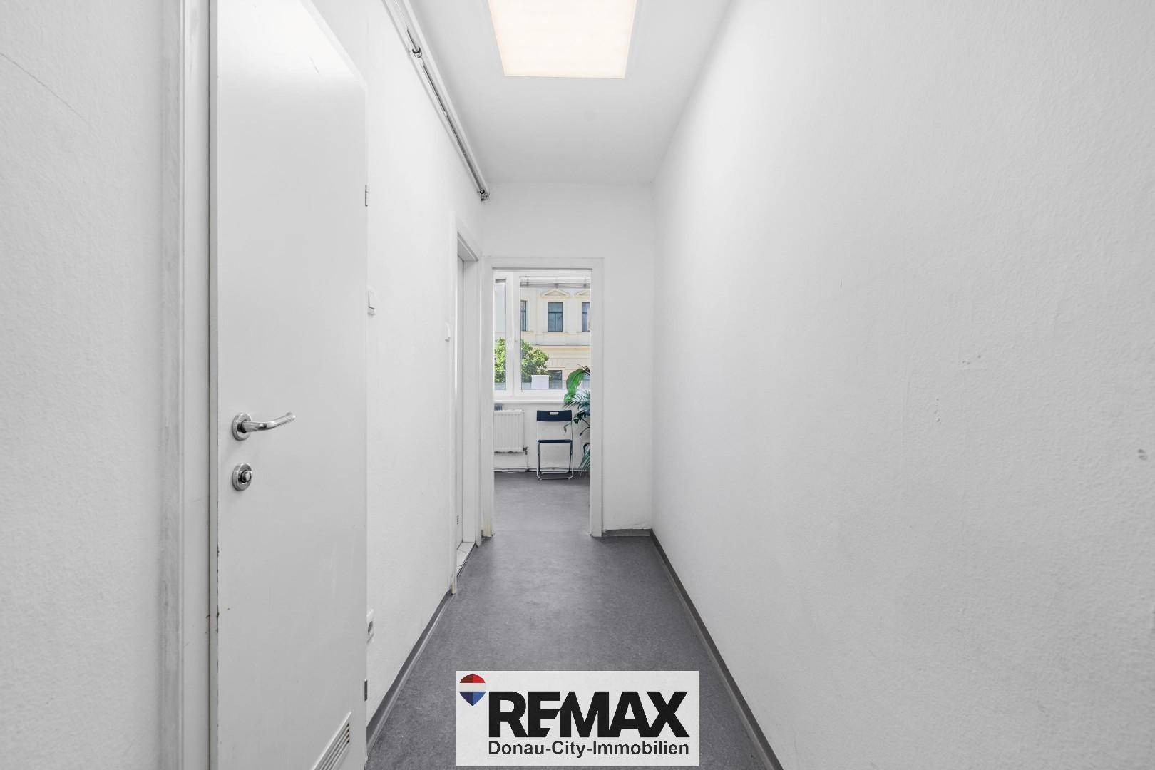Heller Flur mit weißen Wänden und dunklem Boden, führt zu einem Raum mit sichtbarem Fenster.