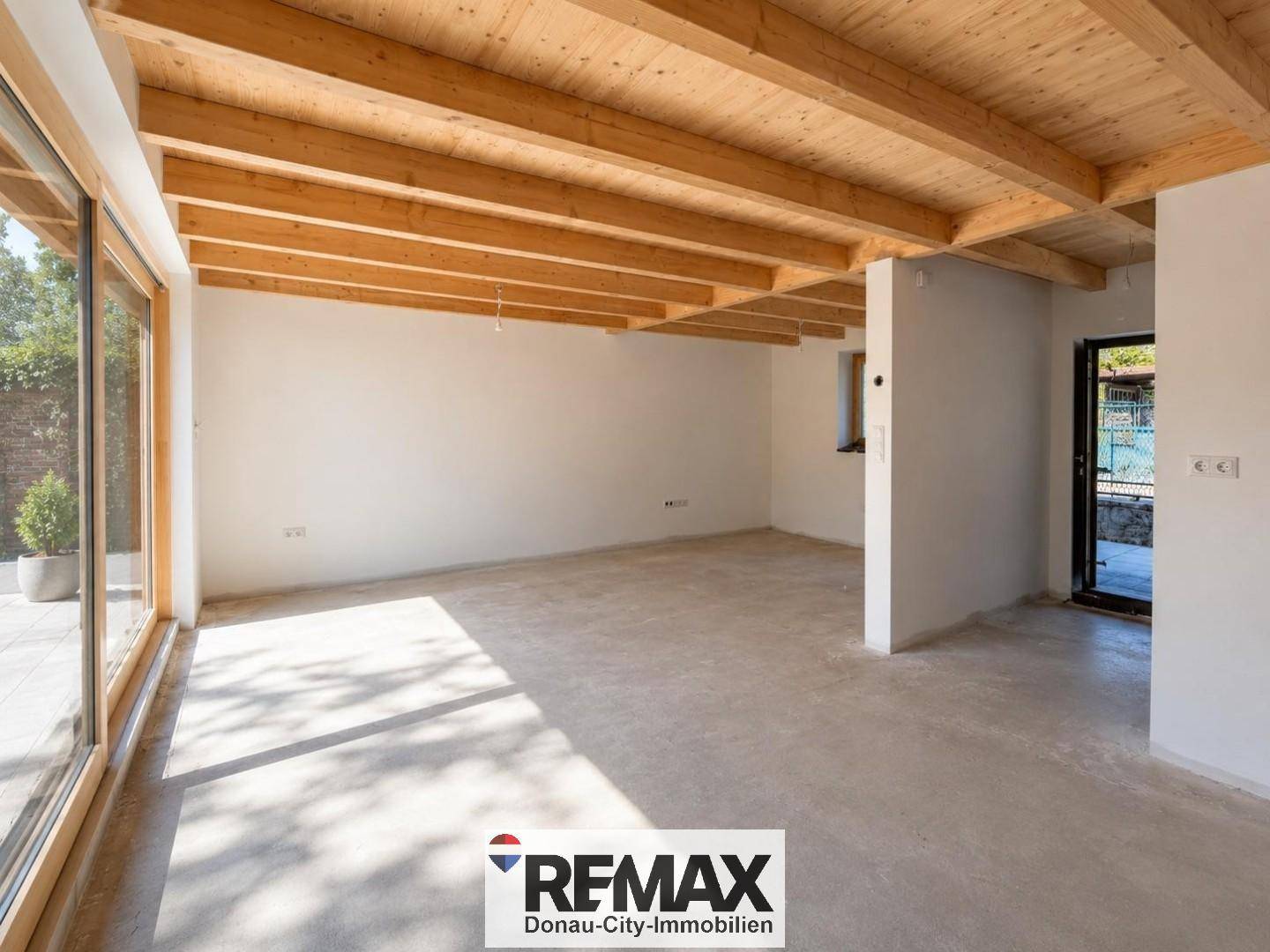Heller Innenraum im Rohbau mit sichtbaren Holzdeckenbalken und großen Glasschiebetüren zum Außenbereich.
